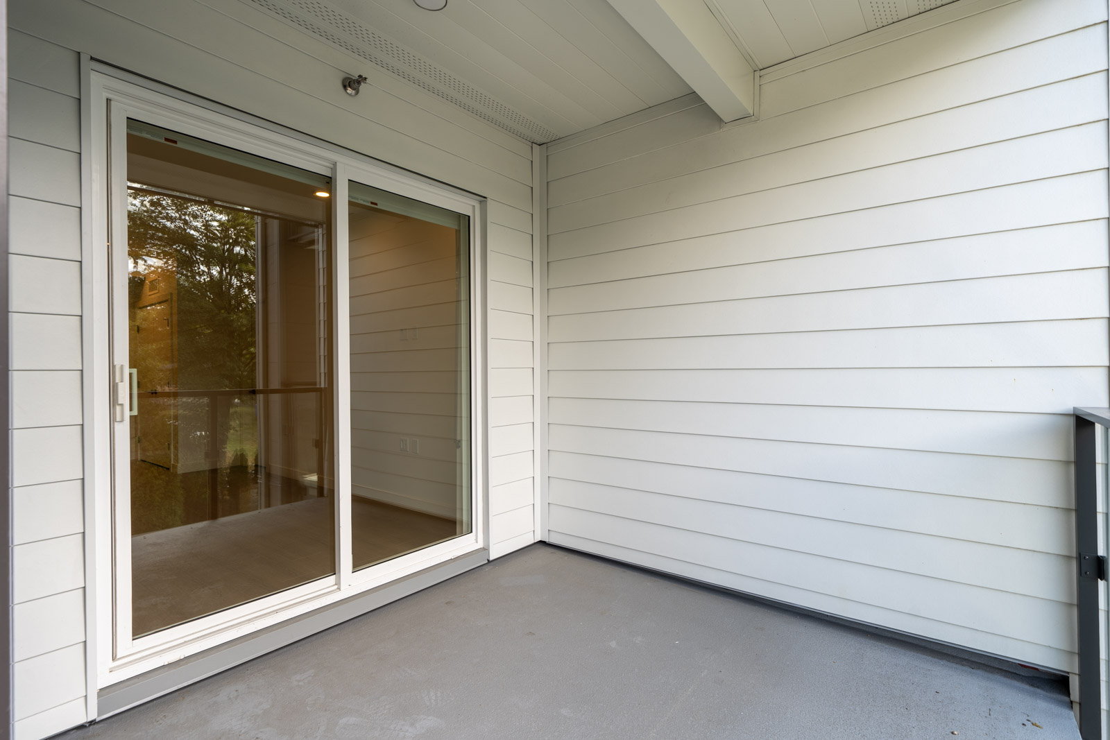Covered balcony with light gray siding, concrete flooring, and a glass sliding door reflecting trees and interior lights.