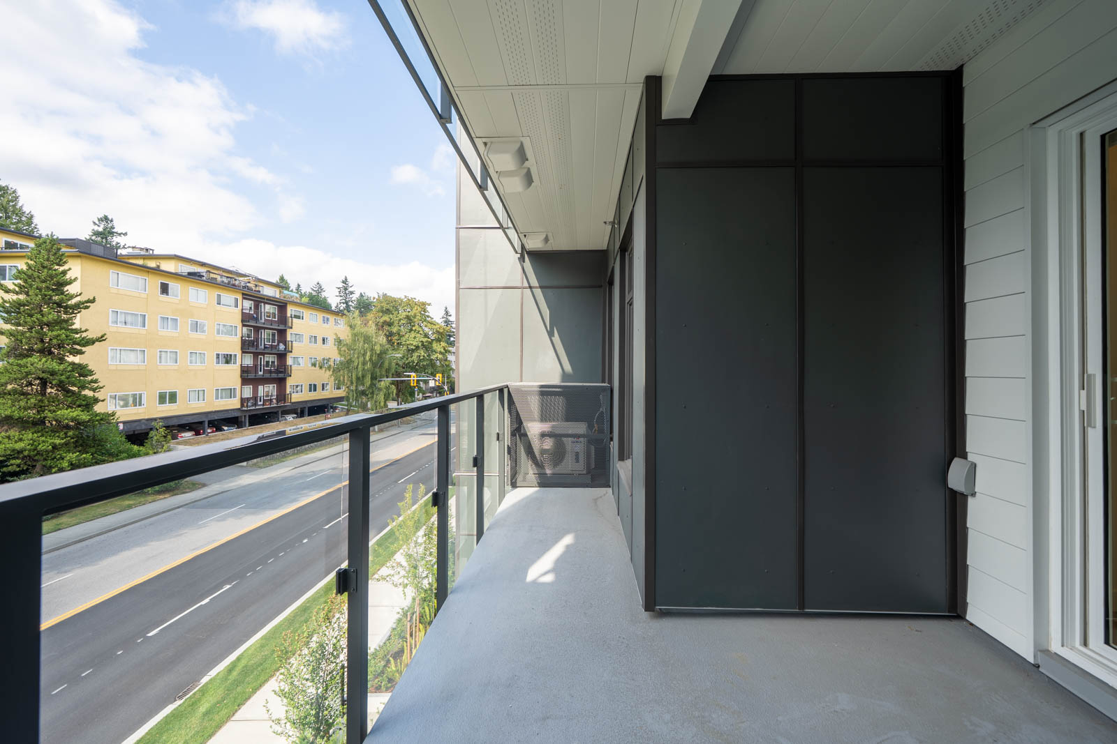 Apartment balcony with glass railing overlooking a street, yellow building and trees in the background.