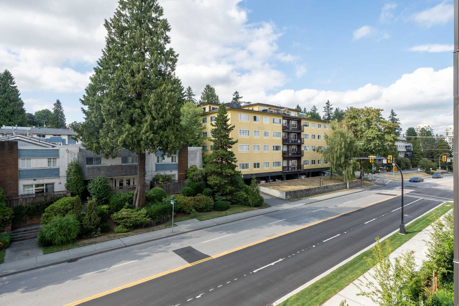 A wide street lined with trees and greenery passes mid-rise apartments under a partly cloudy sky.