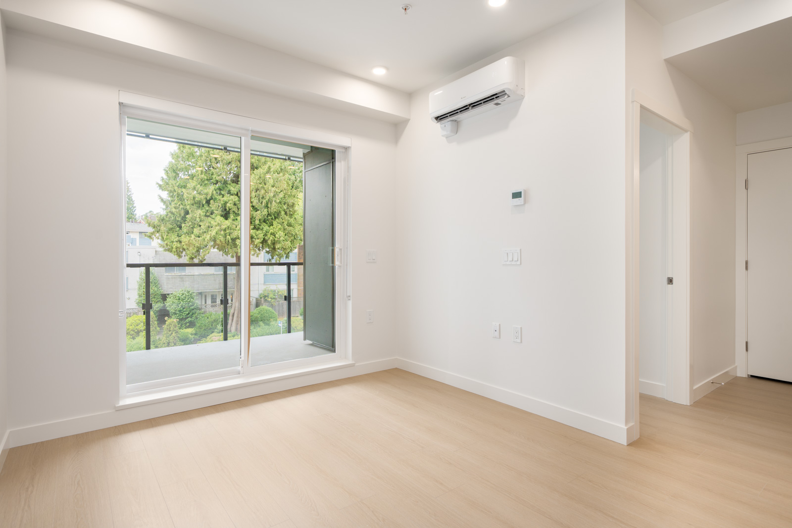 Modern empty room with light wood floors, white walls, sliding glass balcony door, and wall-mounted AC unit.