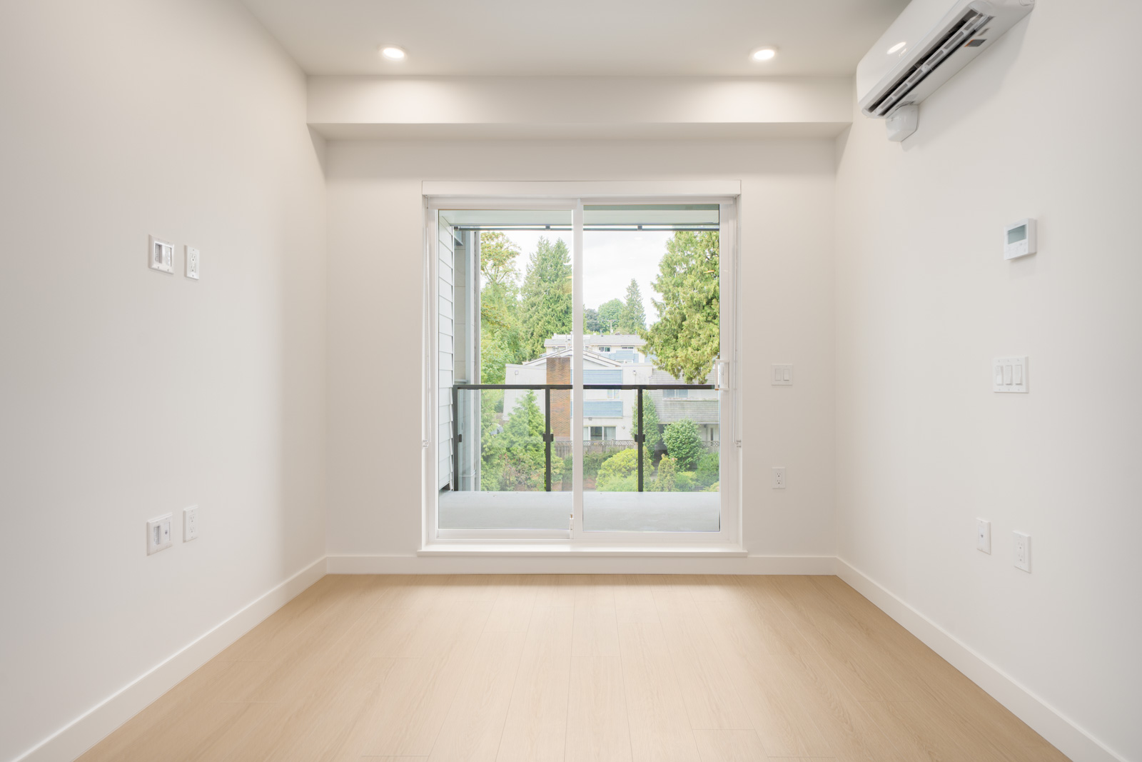 Bright white room with light wood floors, sliding glass doors to a balcony, and a wall-mounted air conditioner.