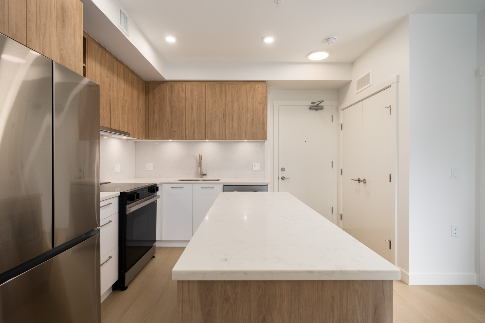Modern kitchen featuring stainless fridge, electric stove, wood cabinets, white counters, island, and light wood floors.
