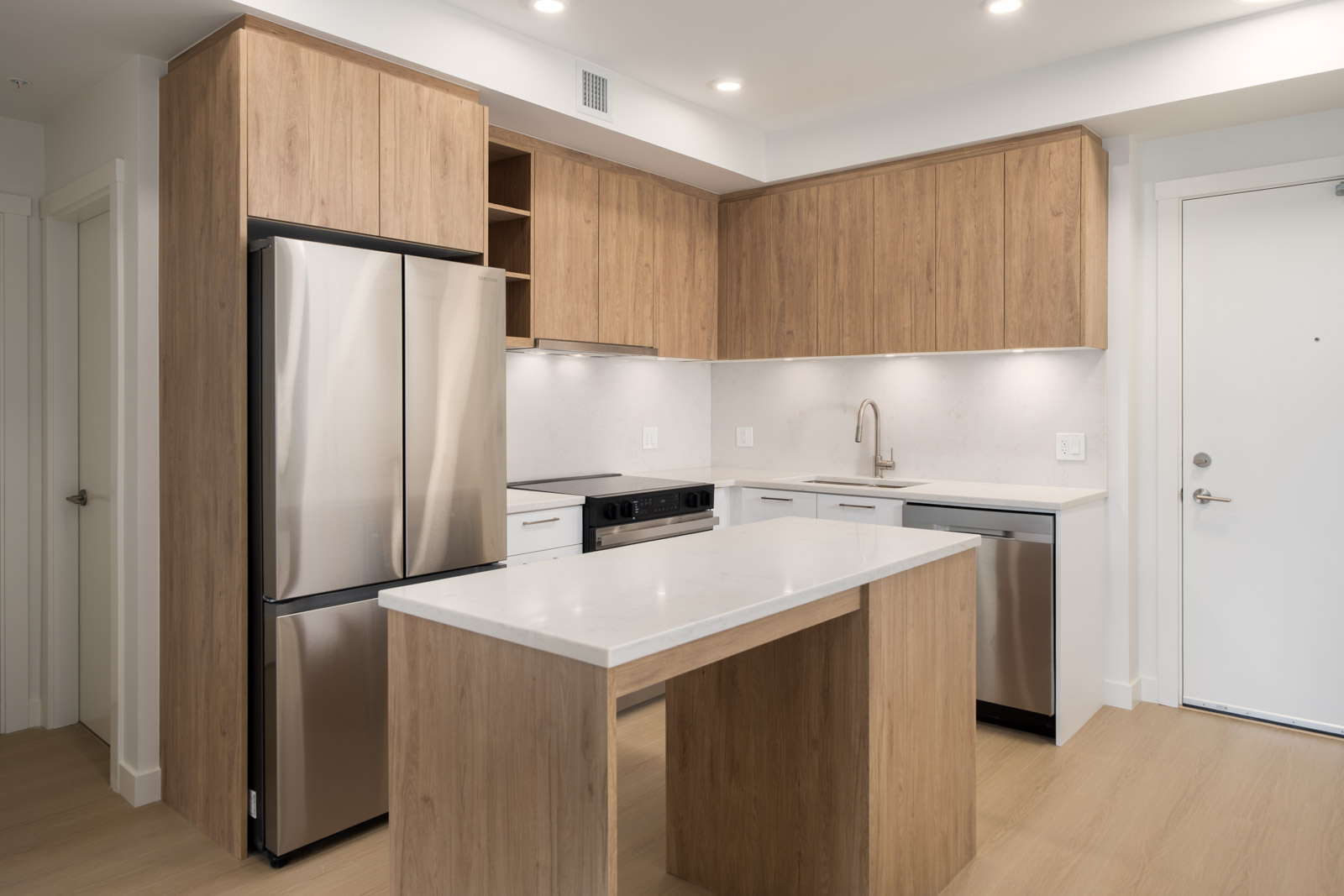 Contemporary kitchen featuring wood cabinets, stainless appliances, white island countertop, and light wood floors under bright lights.