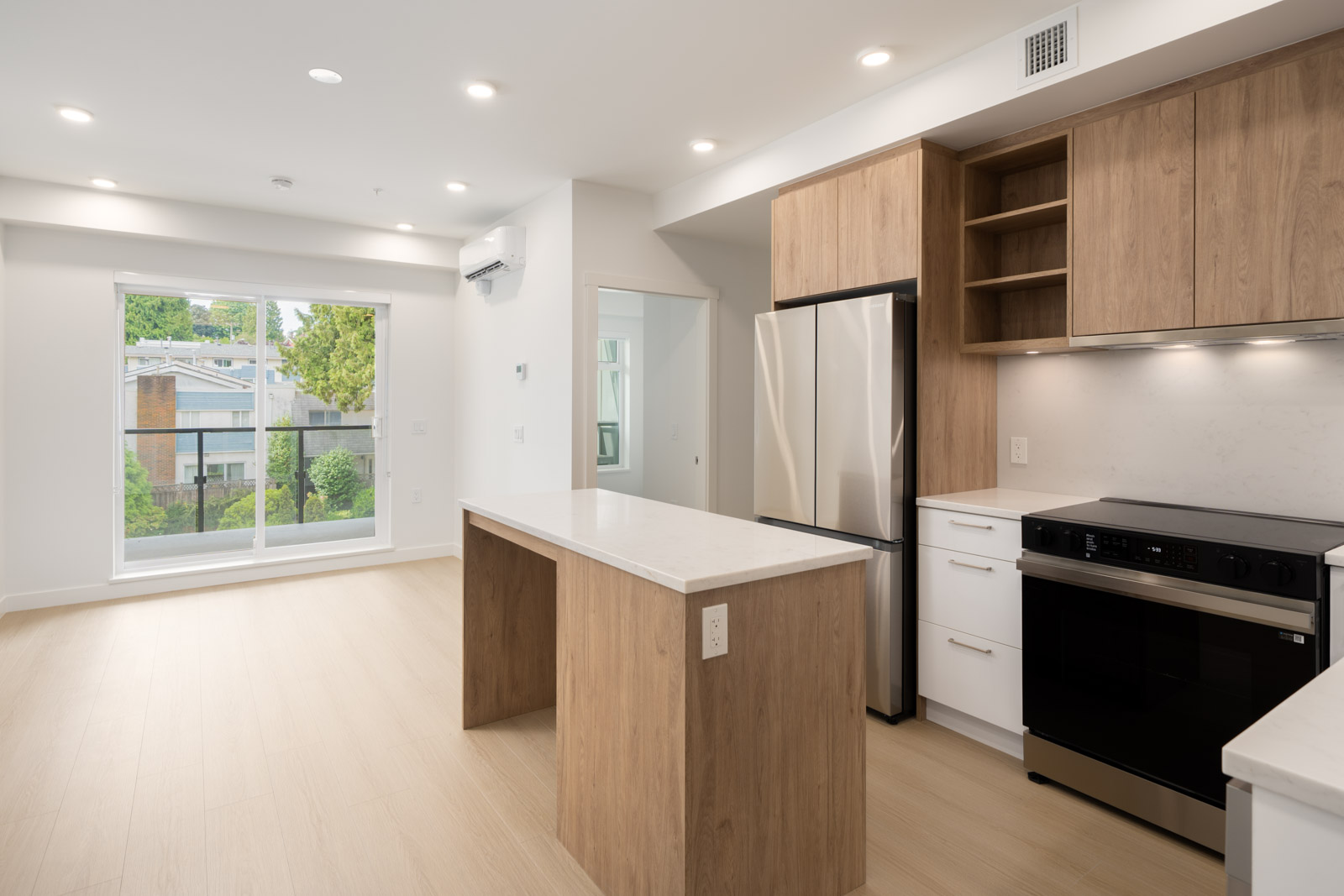 Contemporary kitchen with wood cabinets, stainless appliances, central island, beside a sunny living area with large window and light floors.