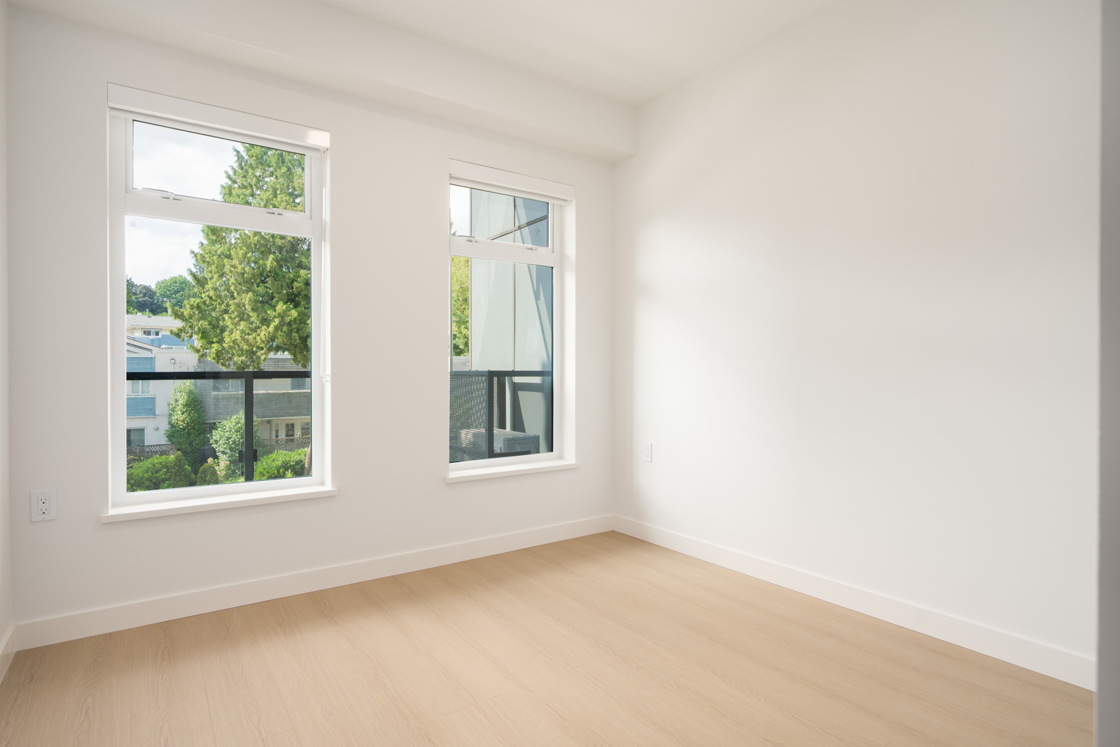Empty room with light wood floors, white walls, and two large windows overlooking trees and nearby buildings.