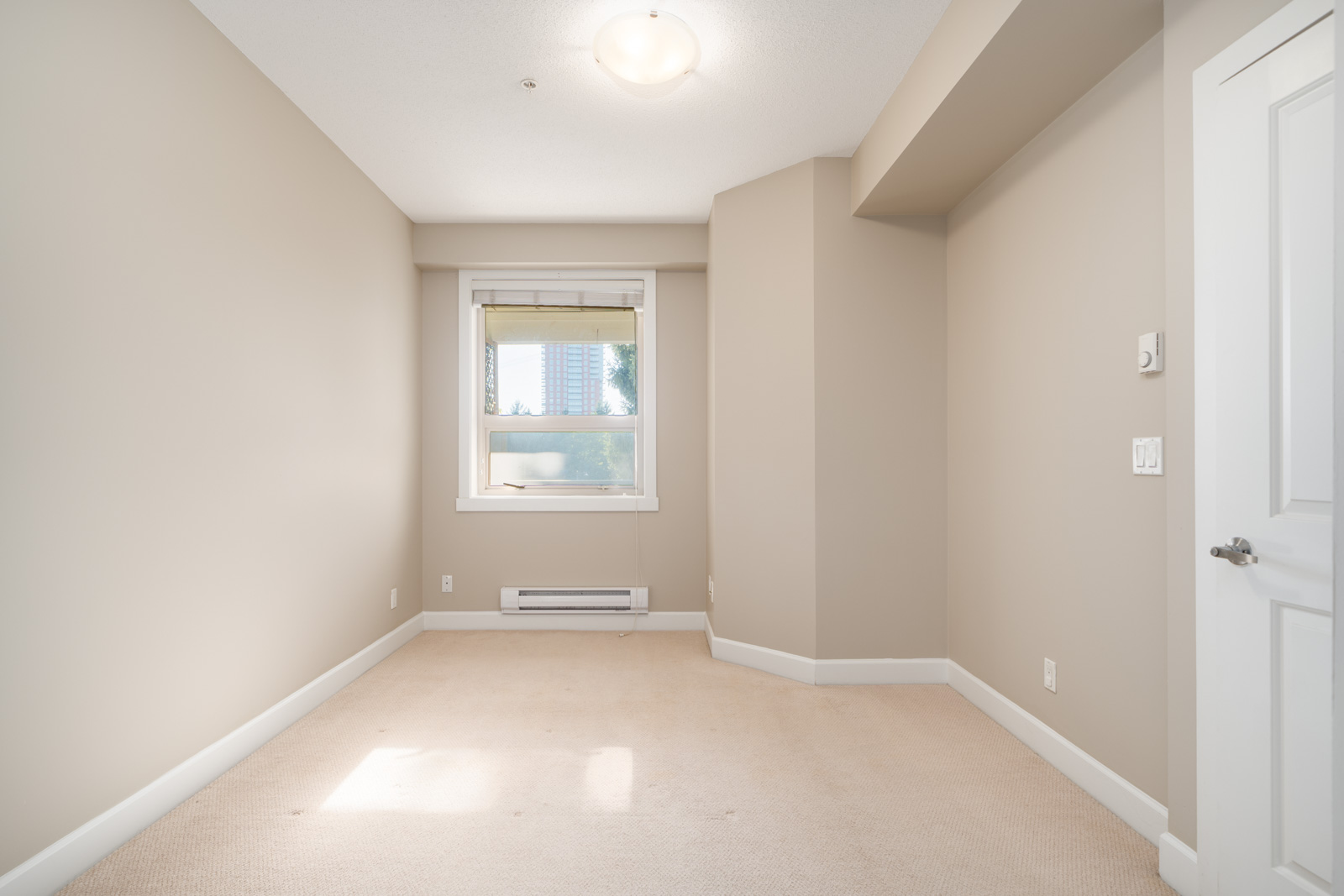 Empty beige-carpeted room with one window, baseboard heater, closed door, and ceiling light; in Surrey at 13897 Fraser Highway.