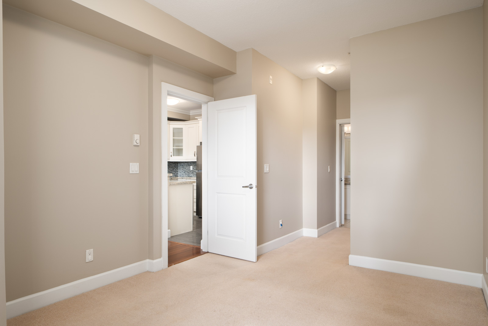 Beige-walled room with light carpet, ceiling light, baseboards; open door to kitchen and hallway with closed doors in Surrey.