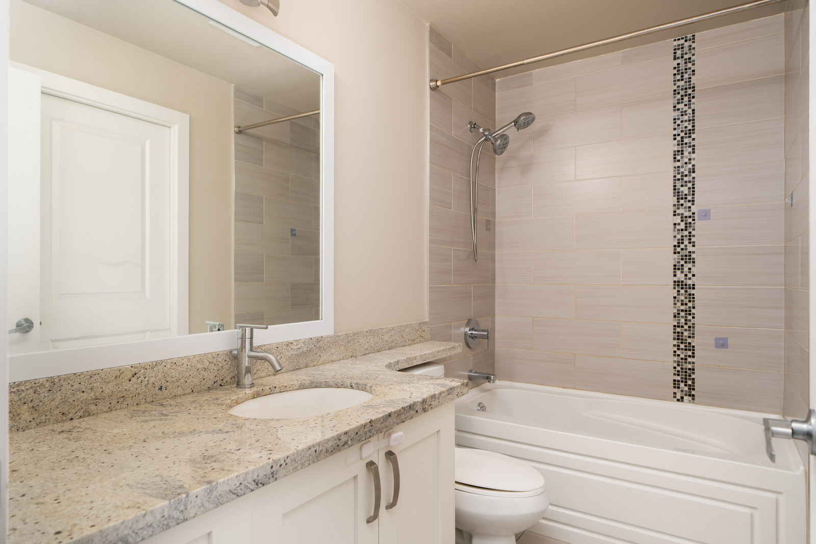 Modern bathroom in Surrey on Fraser Highway with granite countertop, white cabinets, bathtub, and beige tiled walls with mosaic accent.