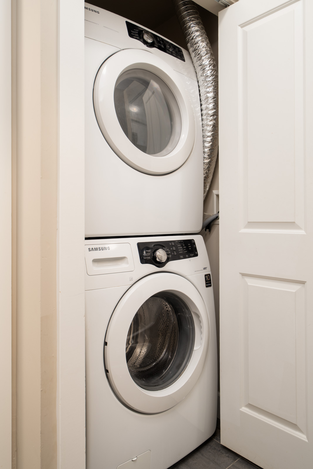 Stacked Samsung washer and dryer in a compact laundry closet with a white door ajar and visible duct, in Surrey near Fraser Highway.