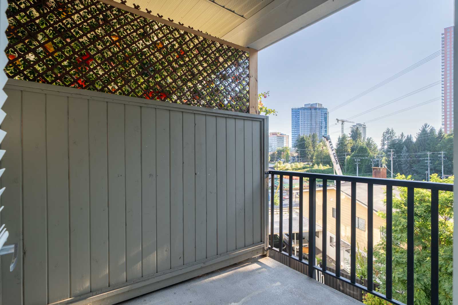 Small balcony with privacy screen, black railing, and partial city view of buildings and trees on a sunny day in Surrey near Fraser Highway.