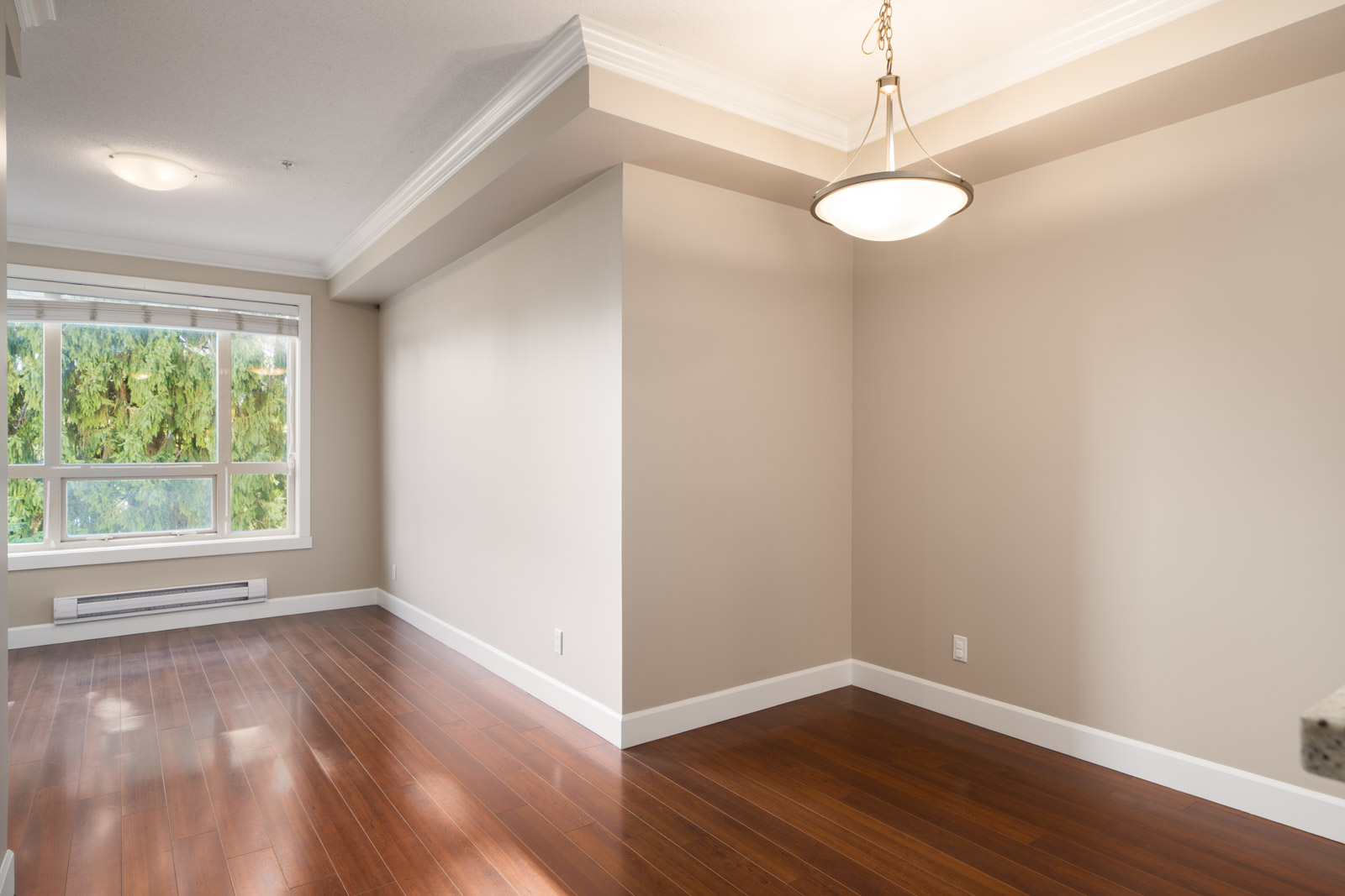 Empty room with hardwood floors, beige walls, large window overlooking trees and Fraser Highway in Surrey, ceiling light above.