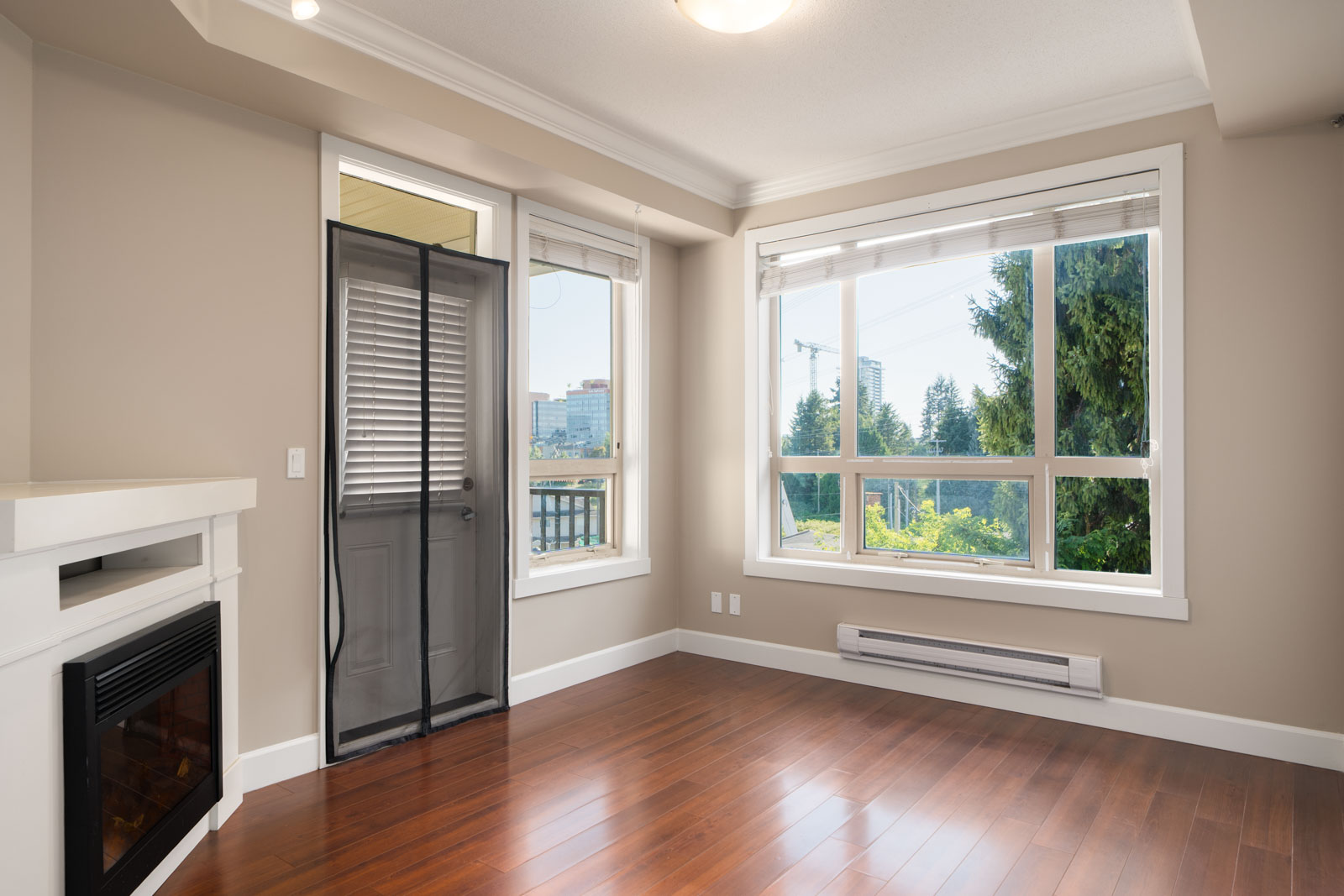 Unfurnished room with wood floors, large windows, a fireplace, and glass door to balcony. Trees and buildings outside in Surrey.