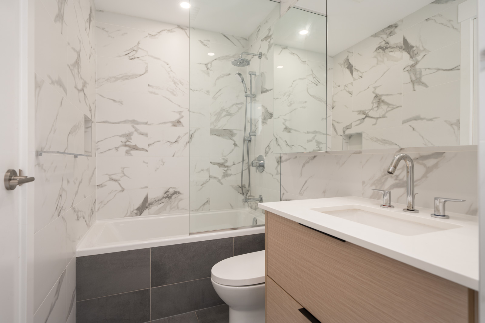 Modern bathroom with marble-patterned walls, glass shower enclosure, gray tile floor, white bathtub, toilet, and a light wood vanity with a white countertop and chrome fixtures.