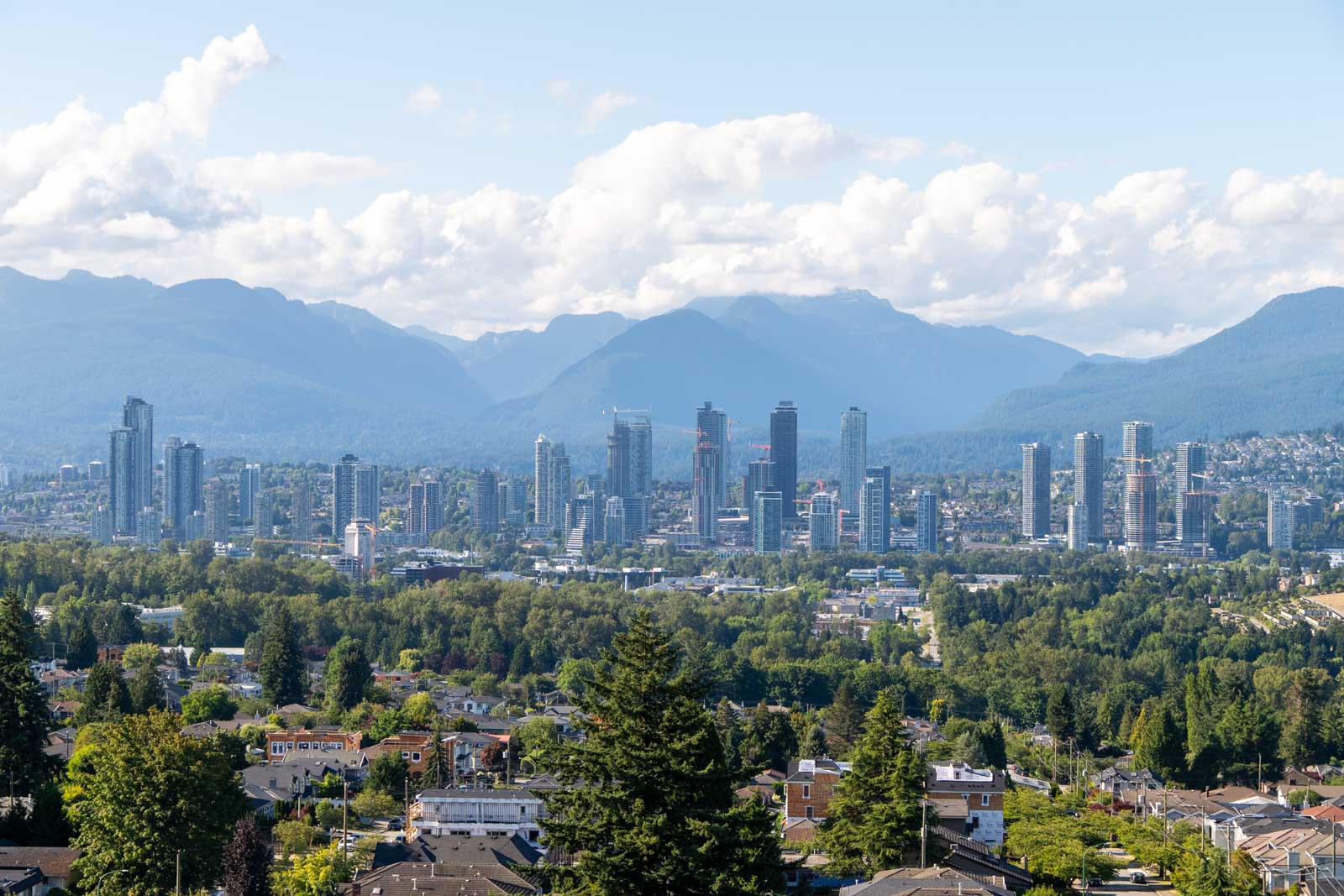City skyline with tall buildings set against a backdrop of mountains, trees, and scattered clouds in the sky.