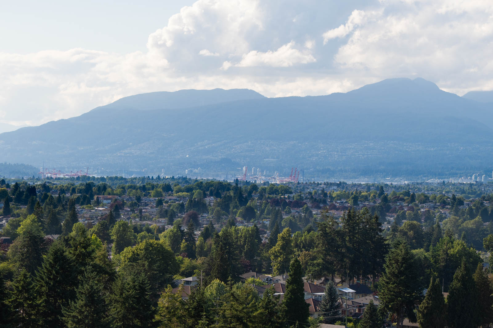 A suburban neighborhood with many trees in the foreground, an industrial area in the middle ground, and mountains under a partly cloudy sky in the background.