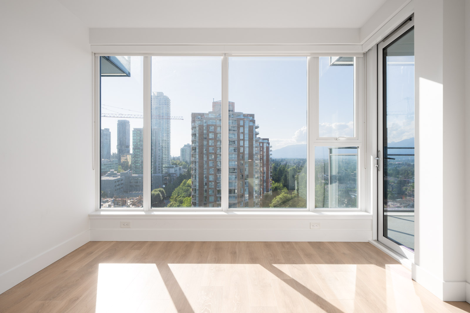 Bright, empty room with light wood flooring and large windows overlooking city buildings and a distant mountain view.