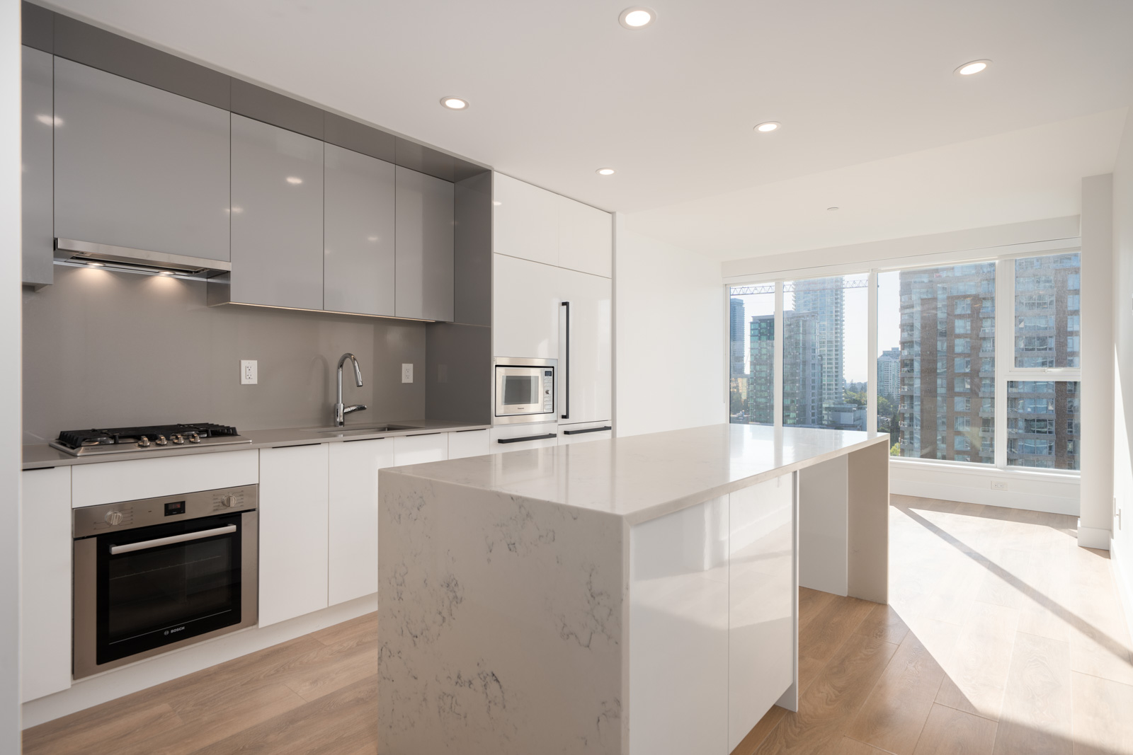 Modern kitchen with glossy cabinets, built-in appliances, a large marble island, wood floors, and tall windows overlooking city buildings in daylight.