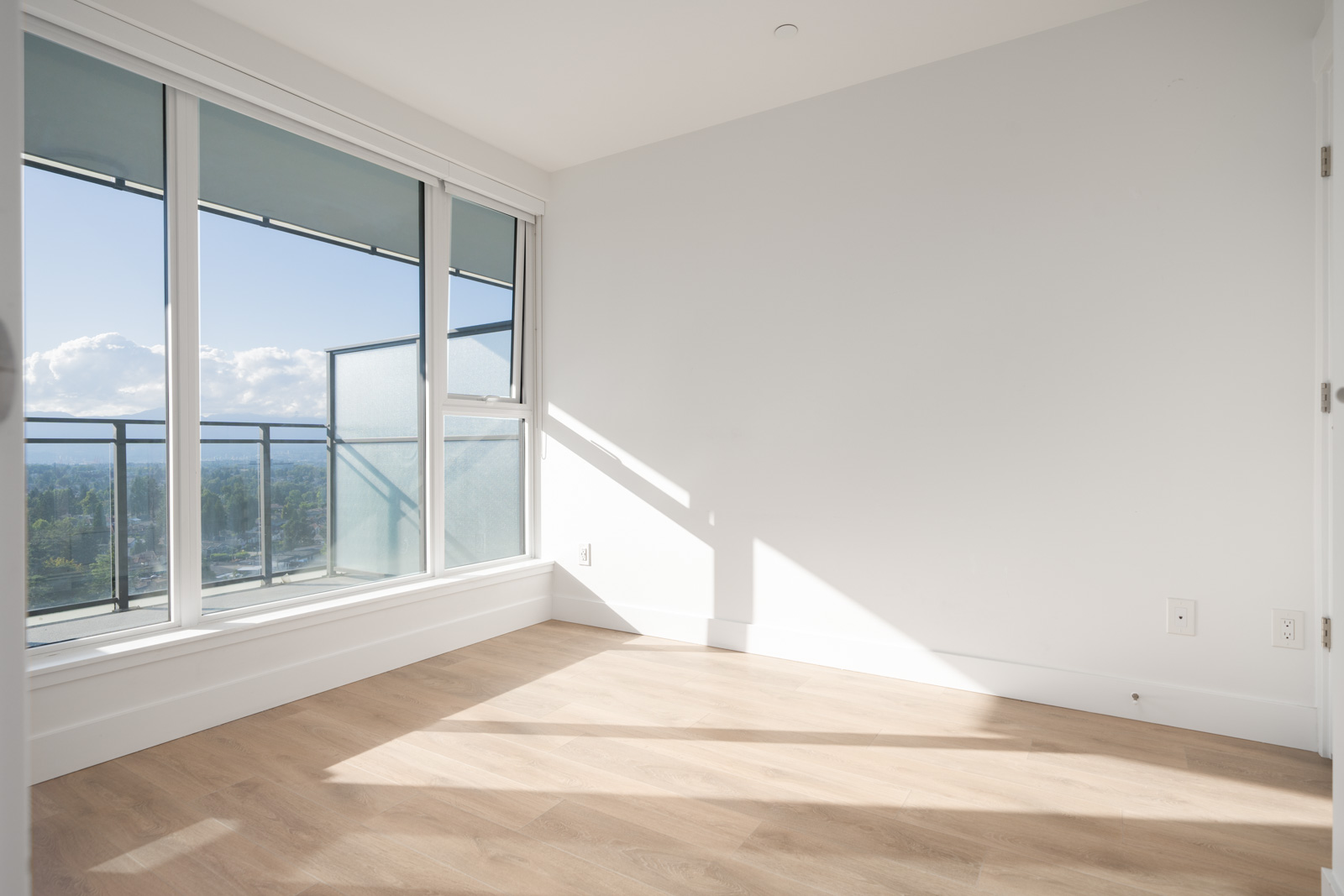 Empty corner room with light wood flooring, white walls, and large floor-to-ceiling windows letting in natural sunlight, overlooking a cityscape.