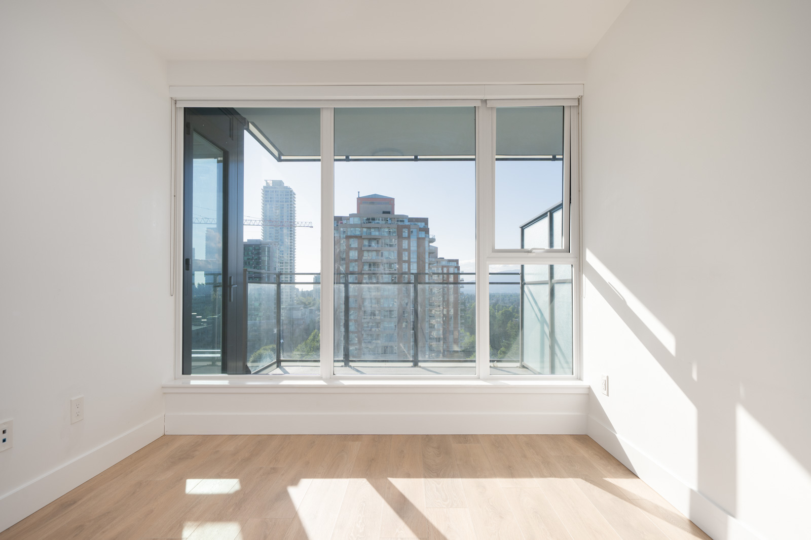 Bright, empty room with light wood floors, white walls, and large windows overlooking a balcony and city buildings. Sunlight casts shadows on the floor.