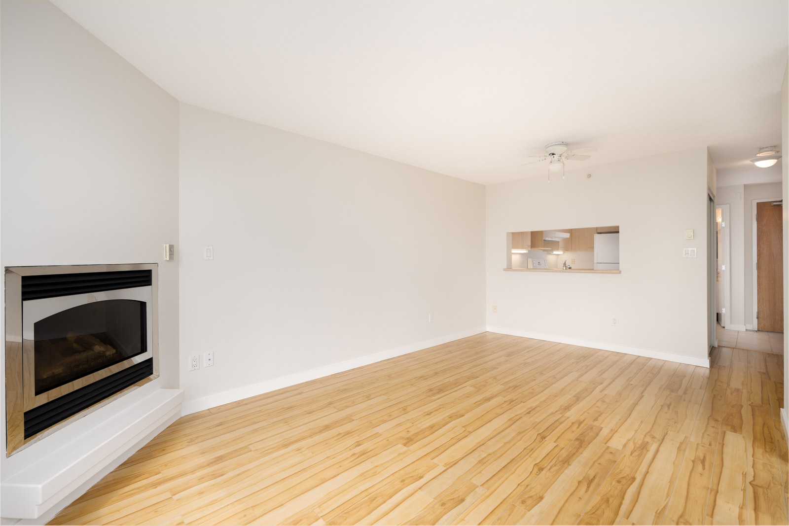 Unfurnished Coquitlam 1st floor living room with light wood floors, corner fireplace, white walls, ceiling fan, and kitchen pass-through.