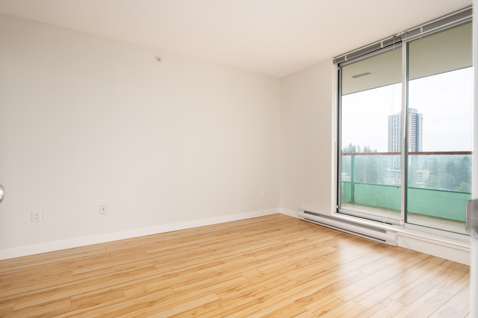 Empty room with light wood floors, white walls, large window, and sliding door to a balcony overlooking Coquitlam city.