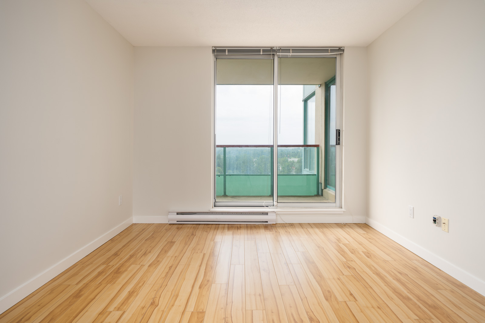 Bright empty room in Coquitlam with light wood floors, white walls, and a sliding glass door to a balcony with green railing.