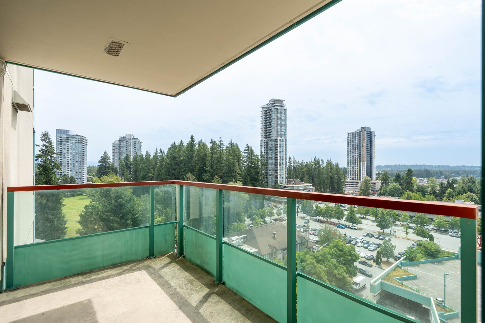 High-rise balcony on Heffley Crescent in Coquitlam with green glass railing, overlooking trees, tall buildings, and sky.