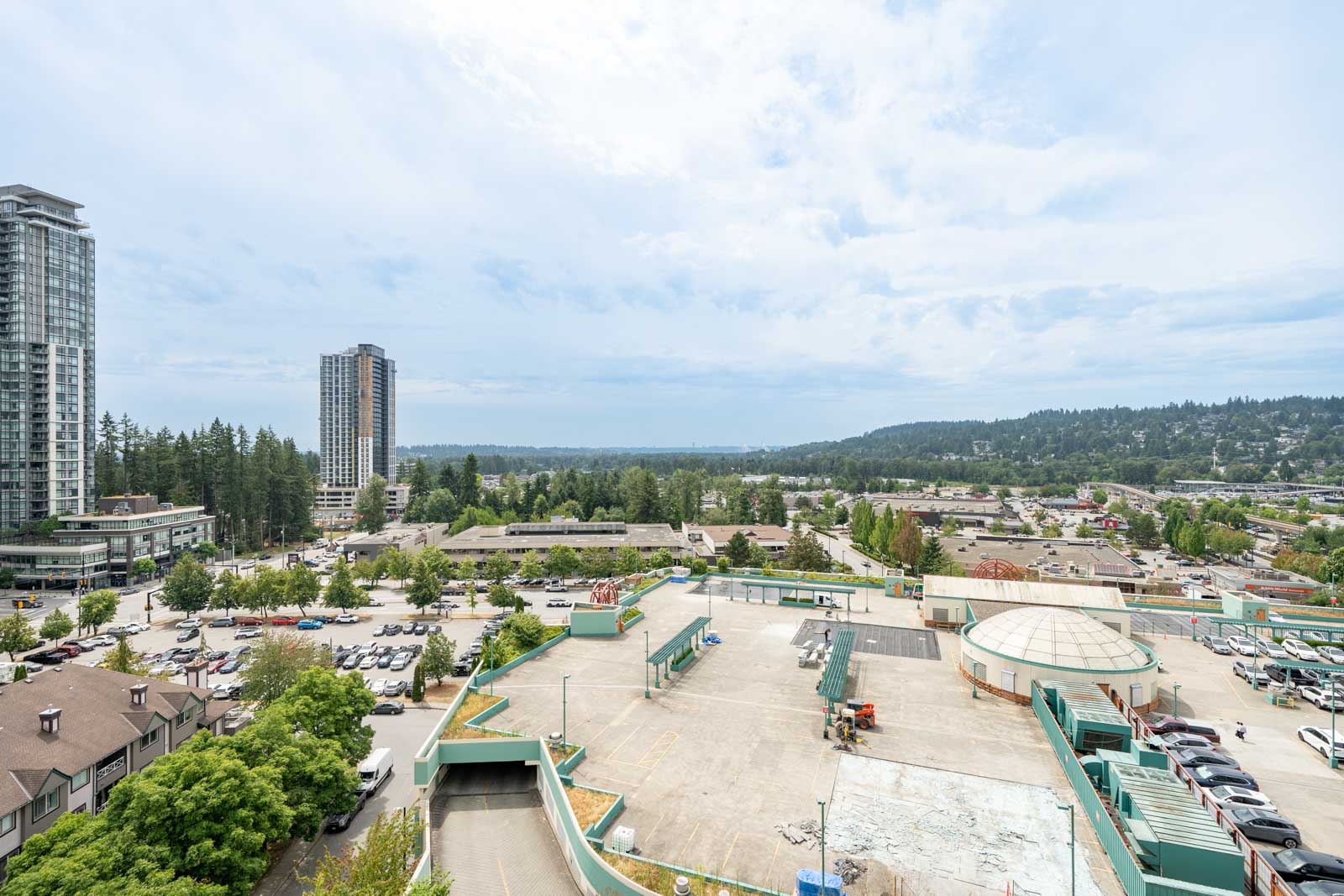 A wide view of Coquitlam, including 1148 Heffley Crescent, with parking lots, towers, trees, and distant hills under clouds.