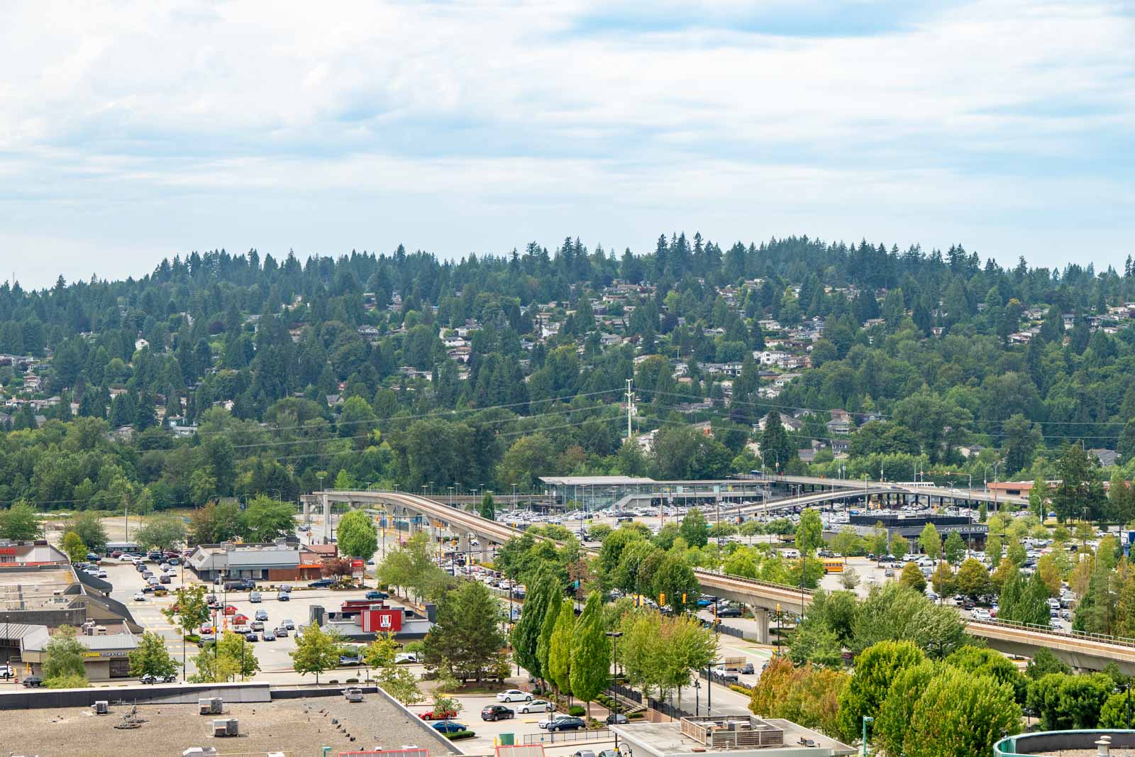 Cityscape of Coquitlam with commercial buildings, traffic, and houses on a tree-covered hillside; Heffley Crescent visible below clouds.