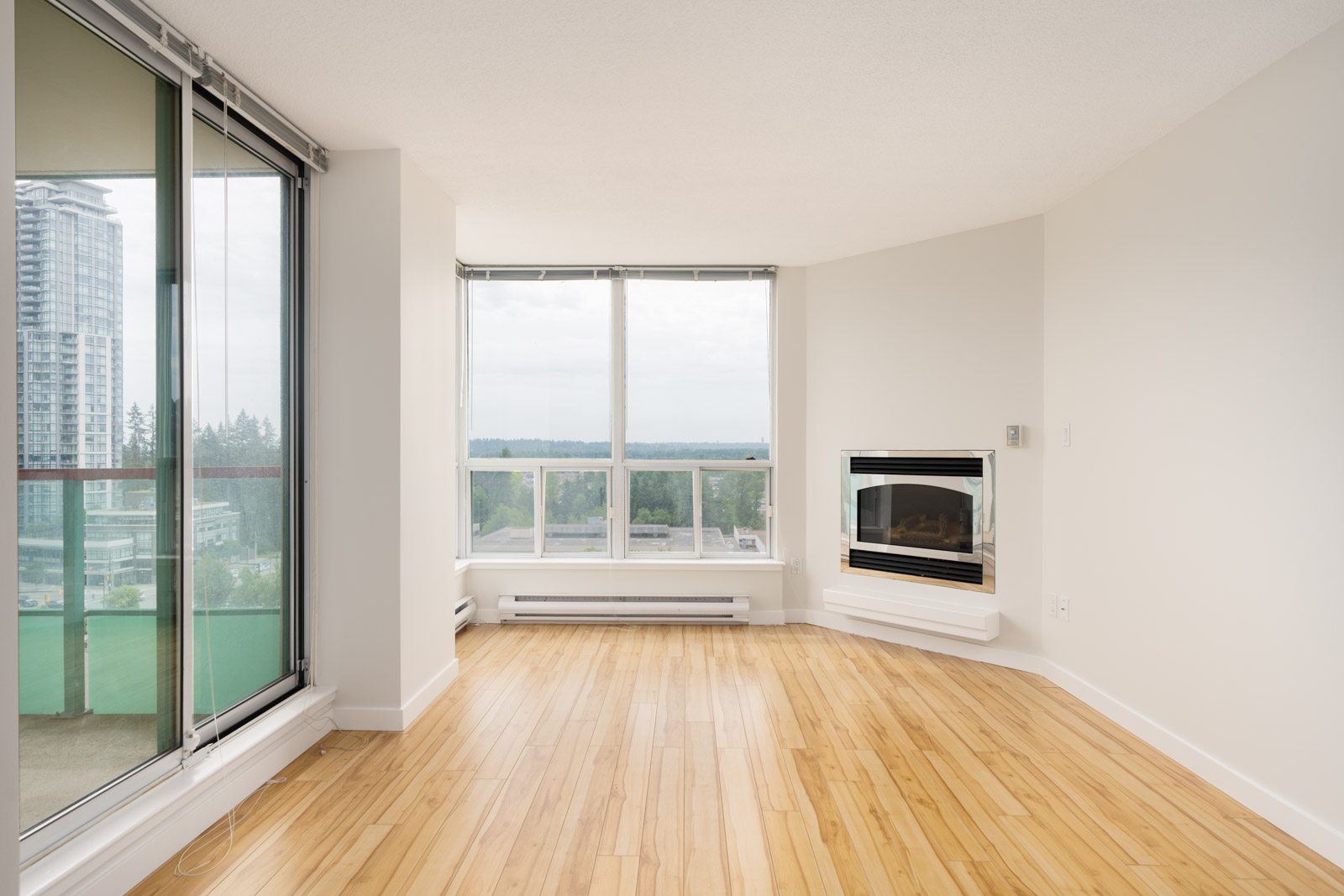 Bright corner apartment room with light wood floors, large windows, glass balcony door, and fireplace, overlooking Coquitlam.