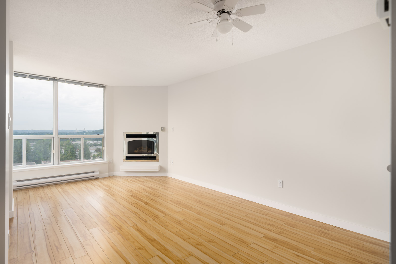 Bright, empty first-floor room with light wood floors, large window, ceiling fan, white walls, corner fireplace in Coquitlam.