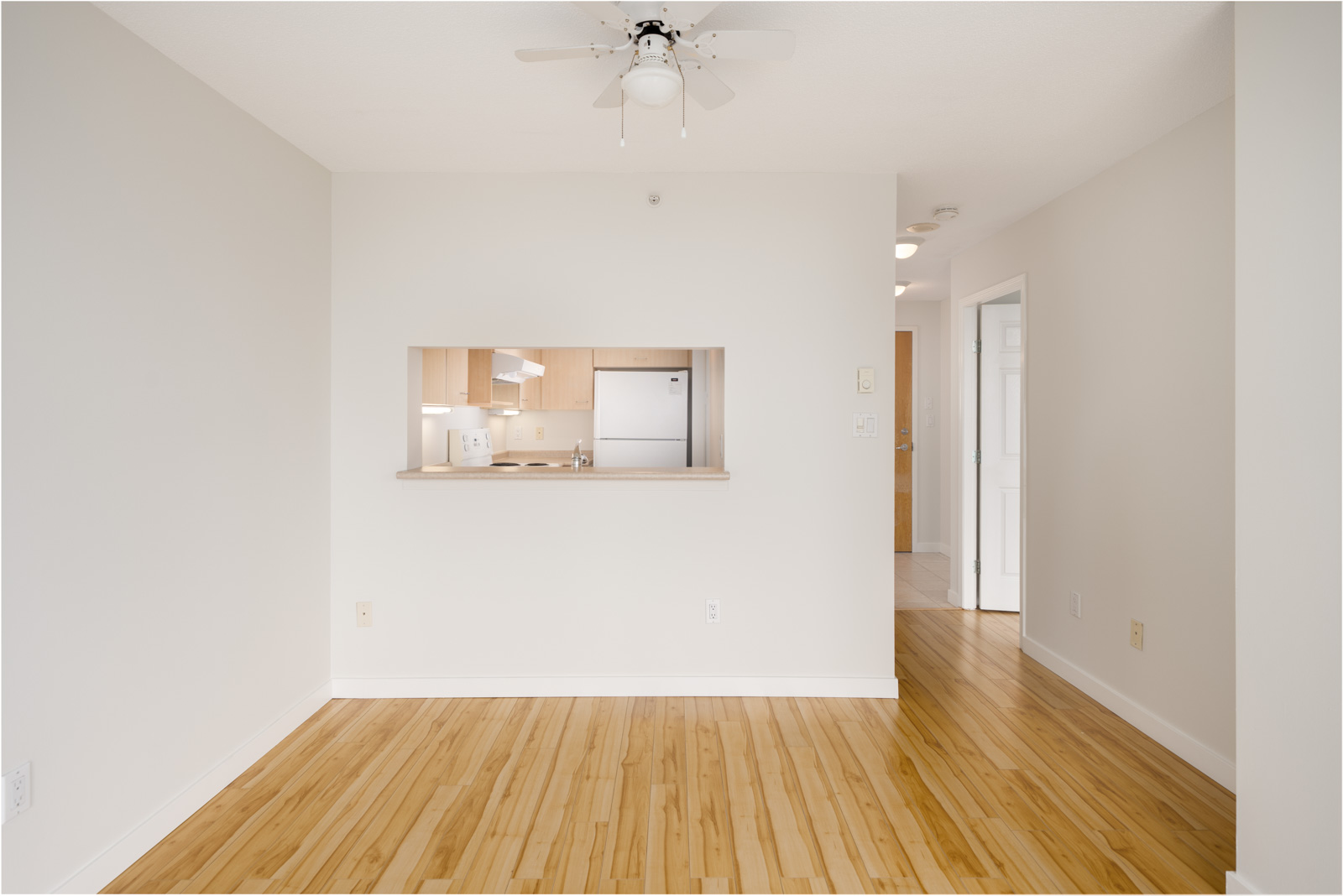 Empty room with light wood floor, white walls, ceiling fan, and a kitchen seen through a pass-through at 1148 Heffley Crescent, Coquitlam.