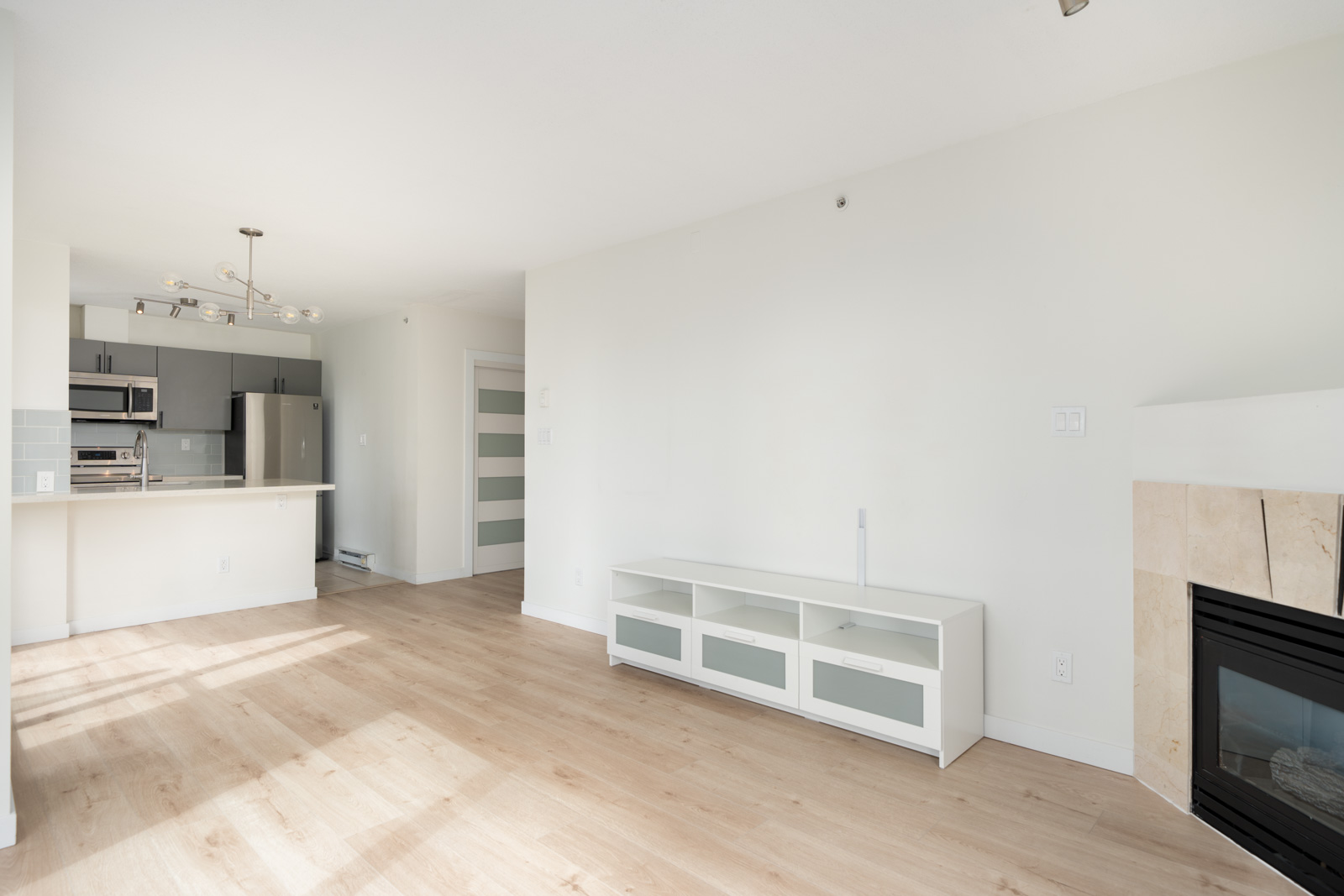 Bright, unfurnished living room with light wood floors, a white TV stand, modern kitchen in the background, and a fireplace on the right.