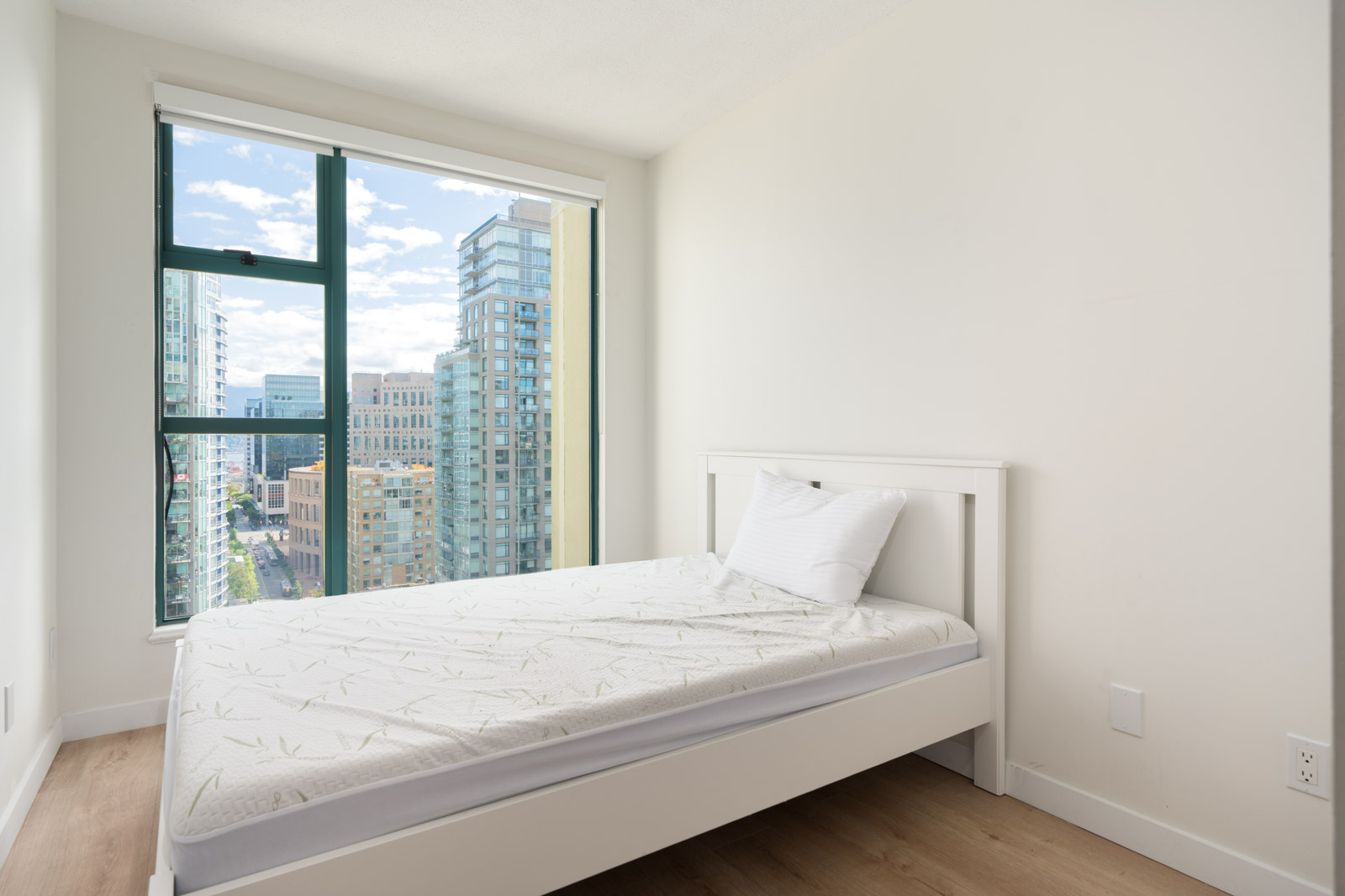 Small bedroom with a single bed, white walls, light wood floor, and a large window showing city buildings under a partly cloudy sky.