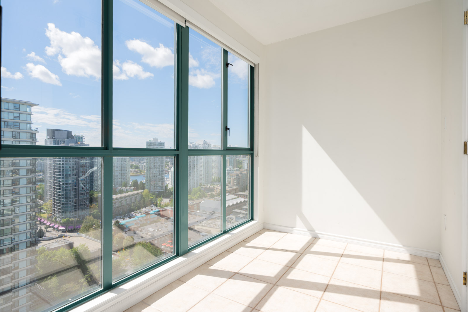 A bright, empty room with large floor-to-ceiling windows offering a city view and sunlight streaming onto the tiled floor.