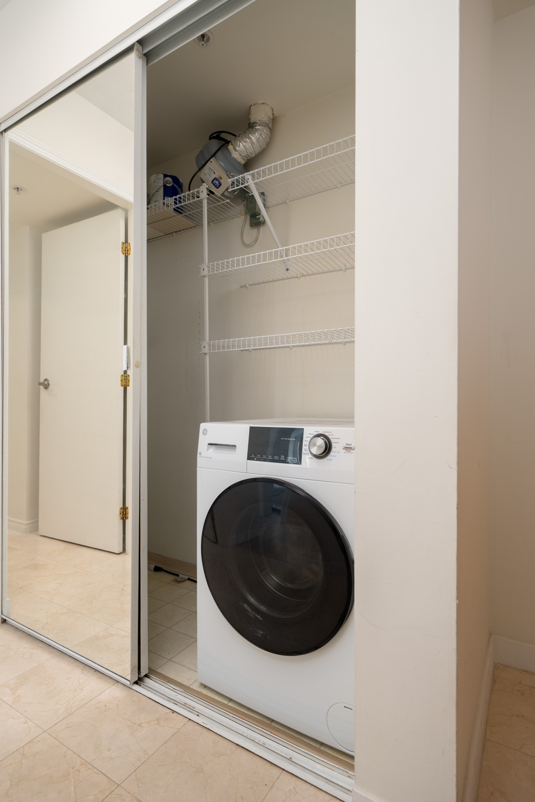 A front-loading washing machine is placed in a small laundry closet with three empty wire shelves and sliding mirrored doors.
