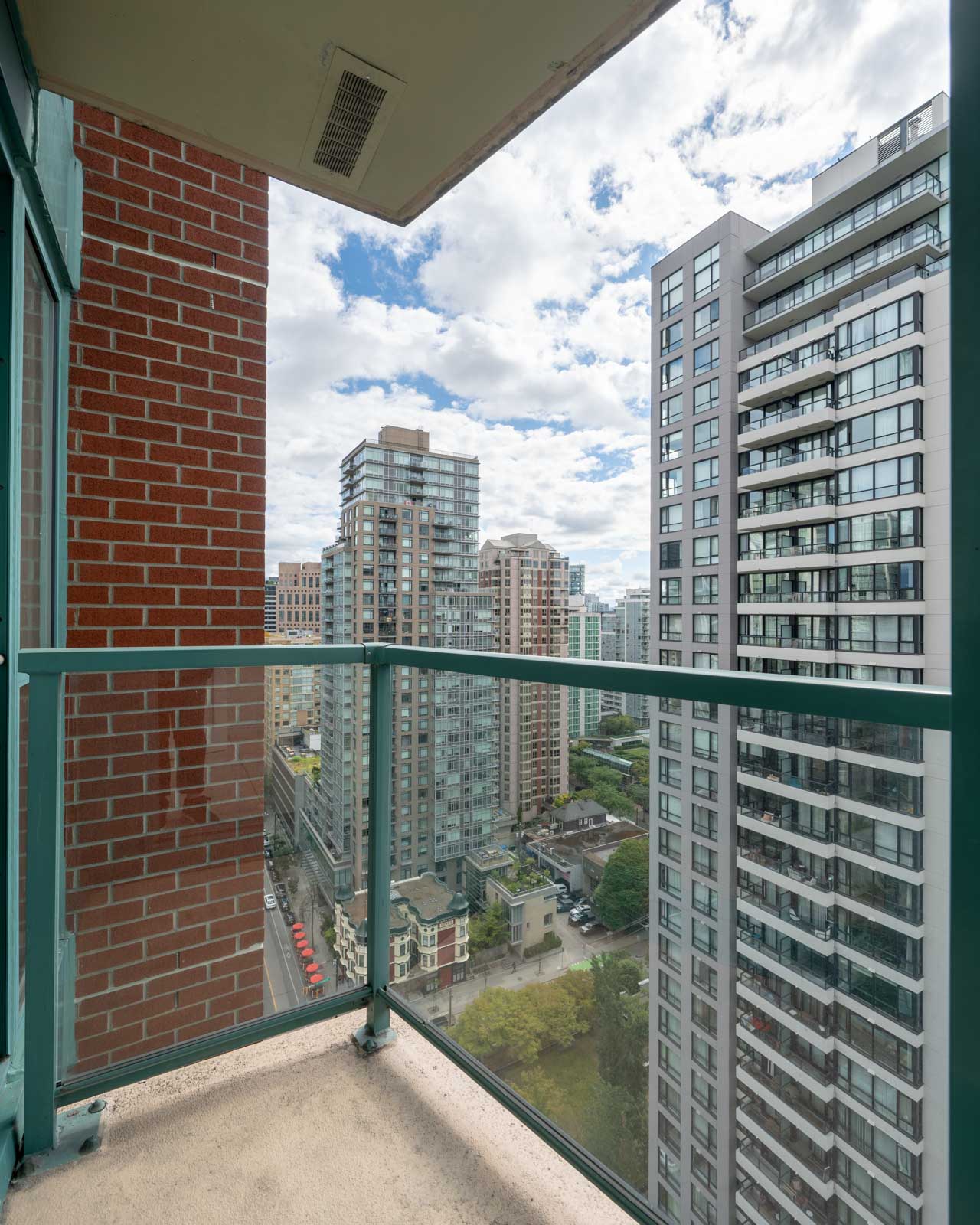 View from a balcony with green railing, overlooking a cityscape of modern high-rise buildings and a street below under a partly cloudy sky.