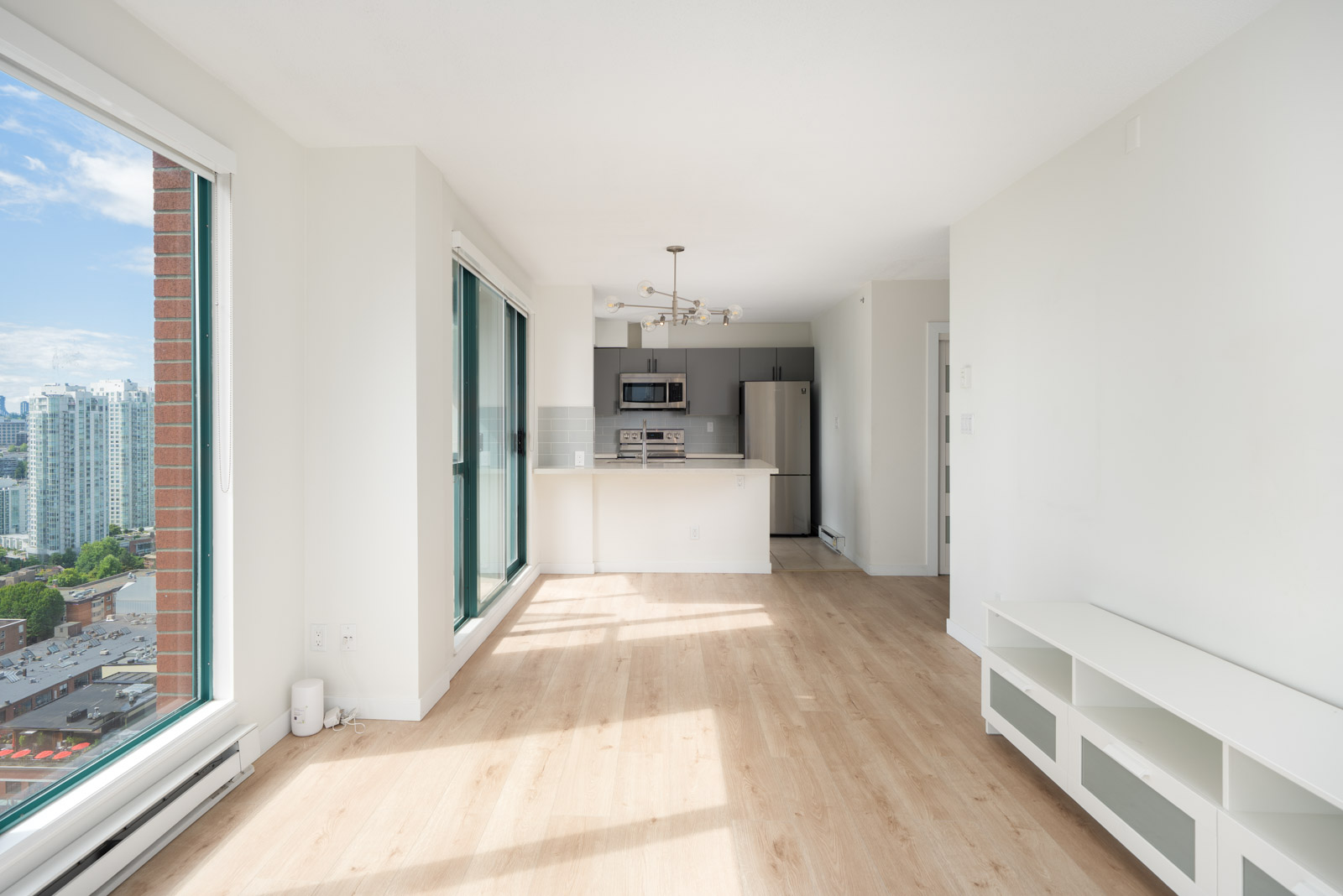 Bright, unfurnished modern apartment with large windows, light wood flooring, white walls, a kitchen area with stainless steel appliances, and a city view.