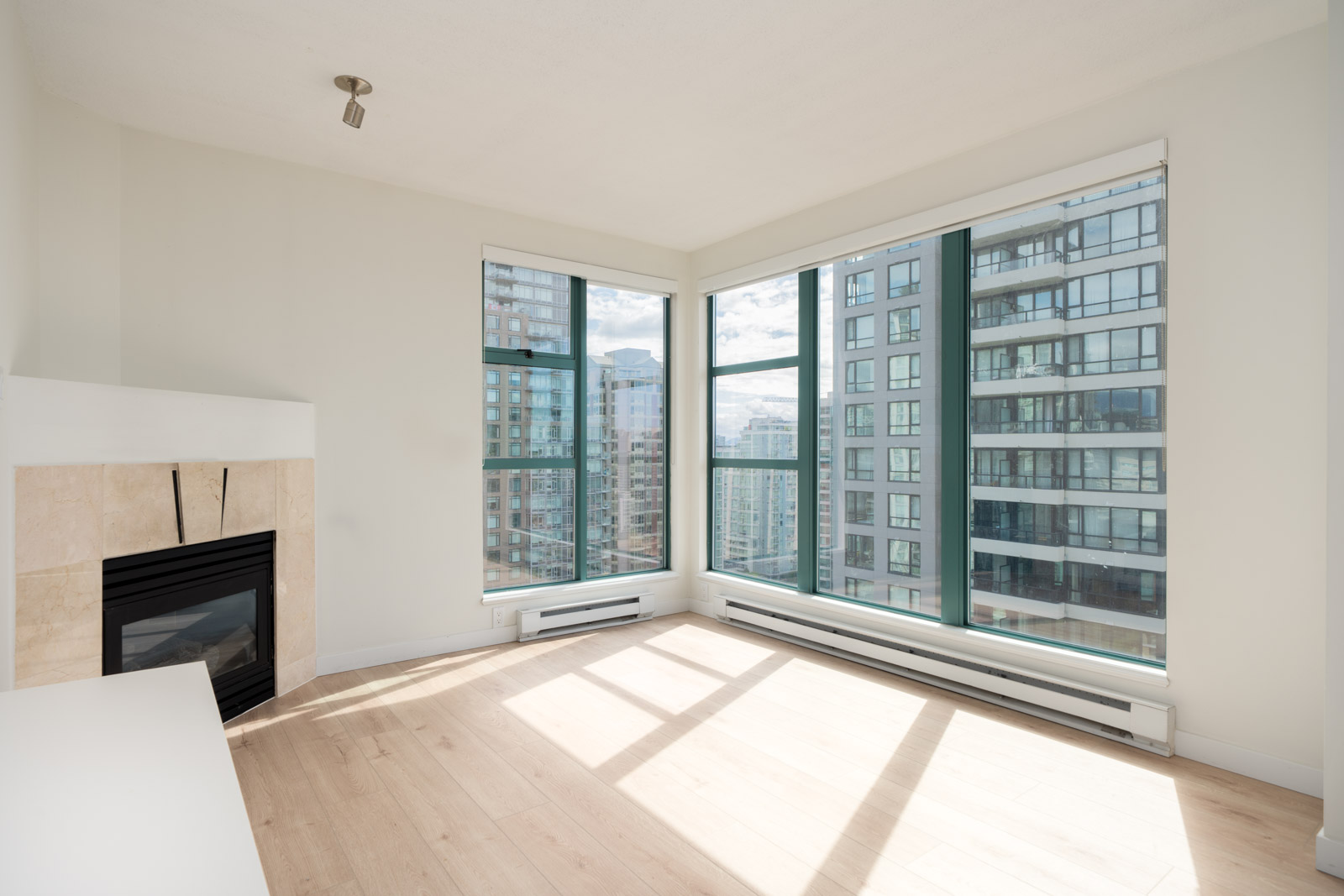 Bright, empty corner room with large floor-to-ceiling windows, light wood flooring, a fireplace, and views of nearby high-rise buildings.