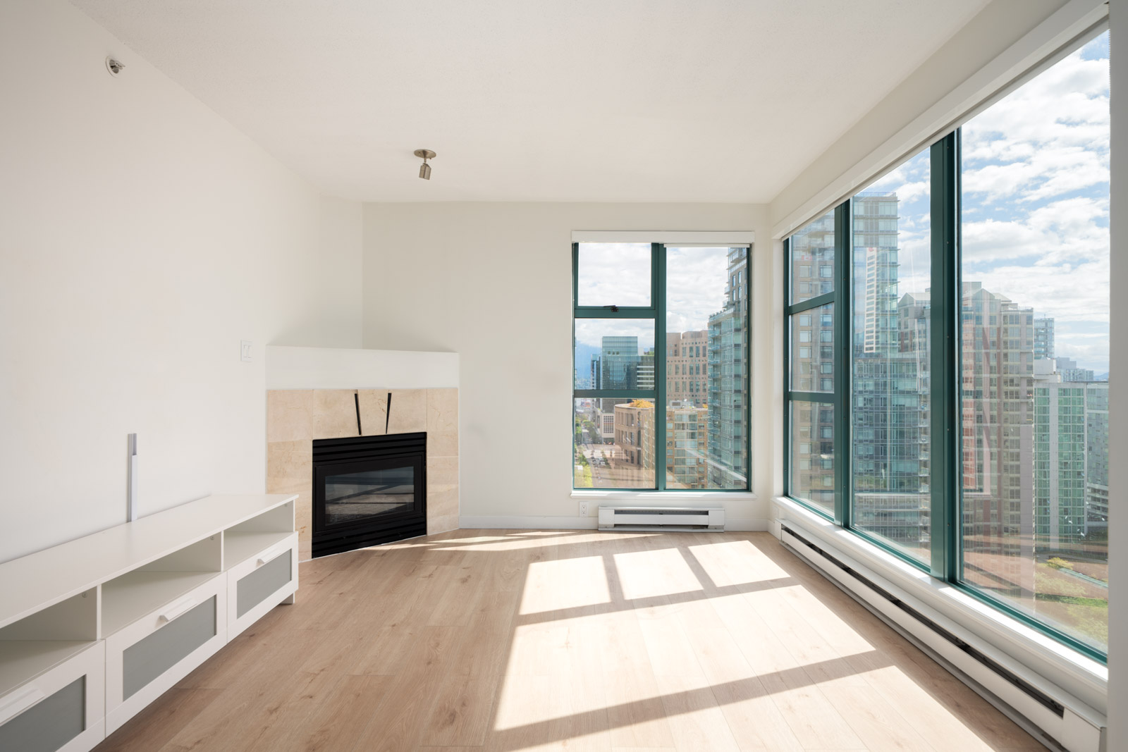 Bright empty corner apartment with large floor-to-ceiling windows, light wood floors, a white media unit, and a modern fireplace overlooking a cityscape.