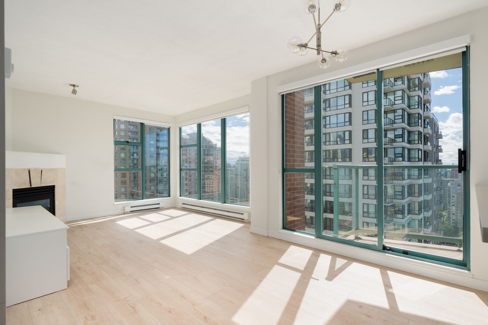 Bright, unfurnished apartment living room with large windows, light wood flooring, modern light fixture, and a partial city view through sliding glass doors and windows.