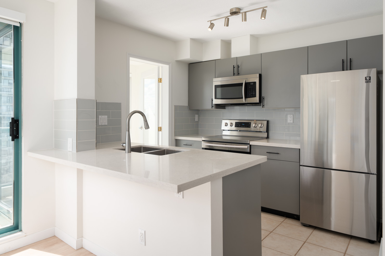 Modern kitchen with gray cabinets, stainless steel appliances, quartz countertop, tile backsplash, and sliding glass door to the left.