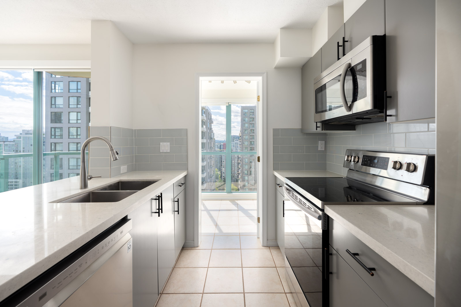 Modern kitchen with gray cabinets, stainless steel appliances, double sink, and large windows showing a city view.