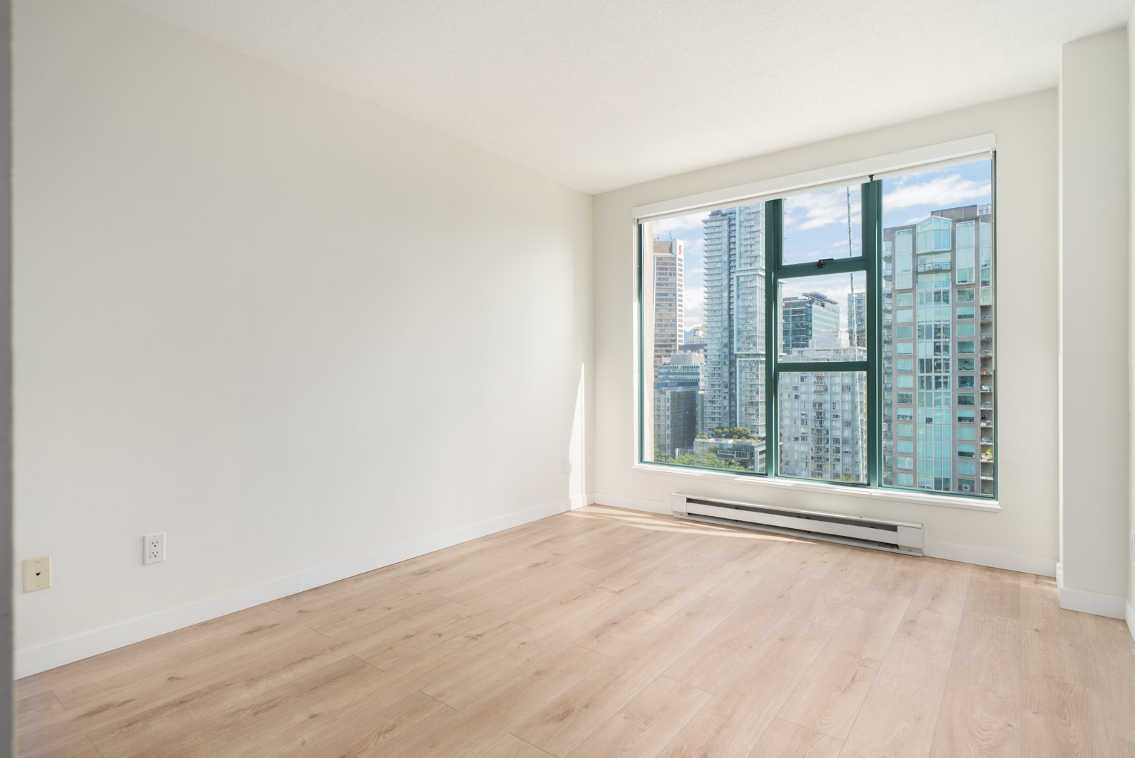 Empty room with light wood flooring, white walls, and a large window showing a cityscape with tall buildings outside.