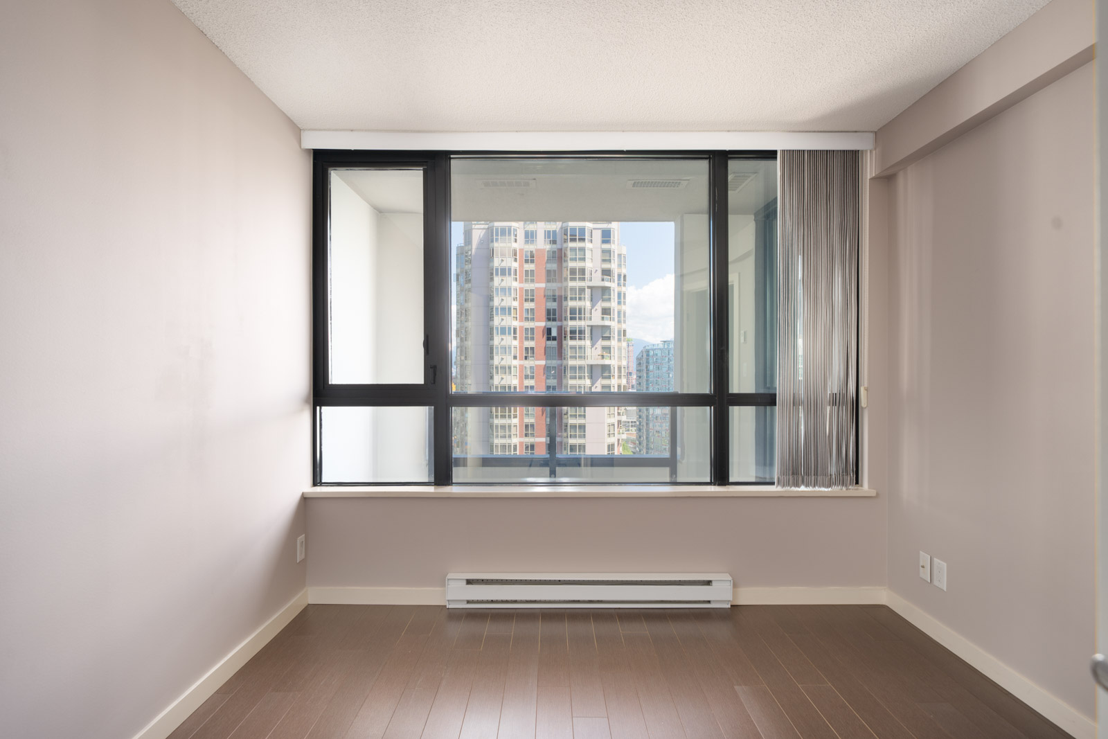 Empty room with light brown walls, wood floors, and a large window overlooking Vancouver city buildings; vertical blinds partly open.