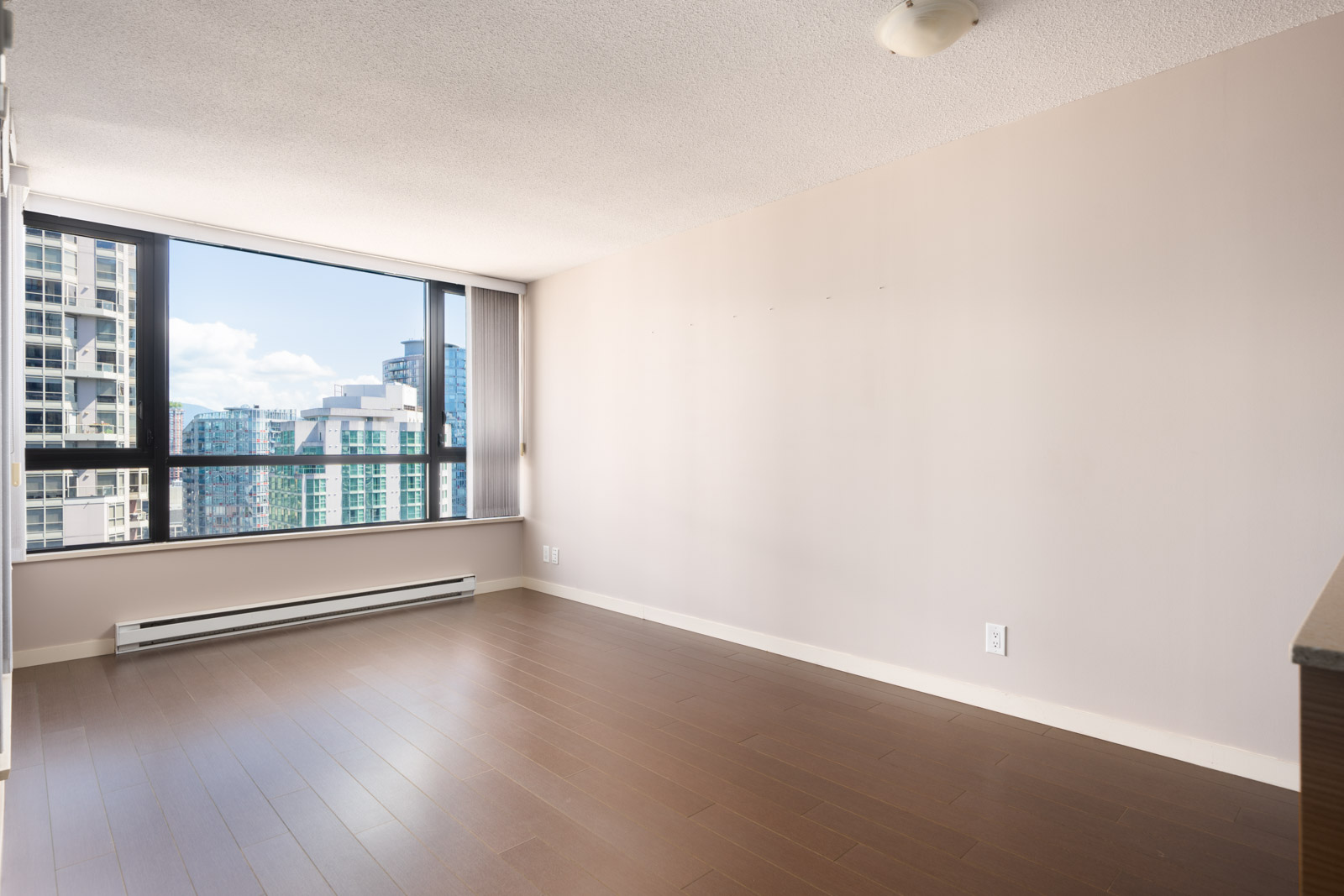 Empty room with light beige walls, large window overlooking Vancouver city buildings, brown wood floor, and ceiling light fixture.