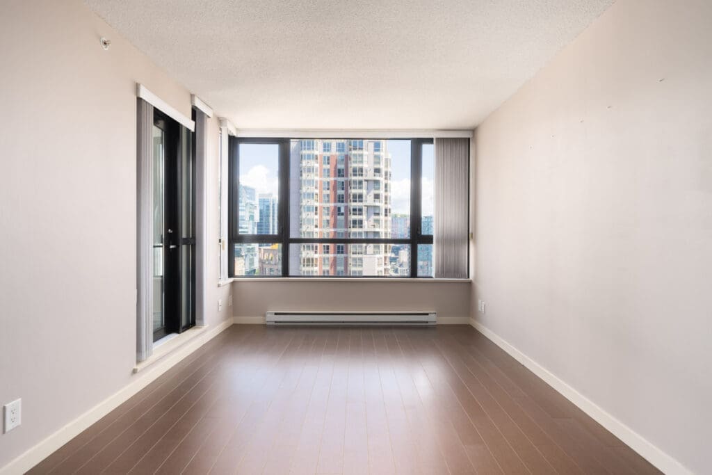 Empty apartment with light wood floors, beige walls, large window overlooking Vancouver from the 23rd floor, and a glass door left.