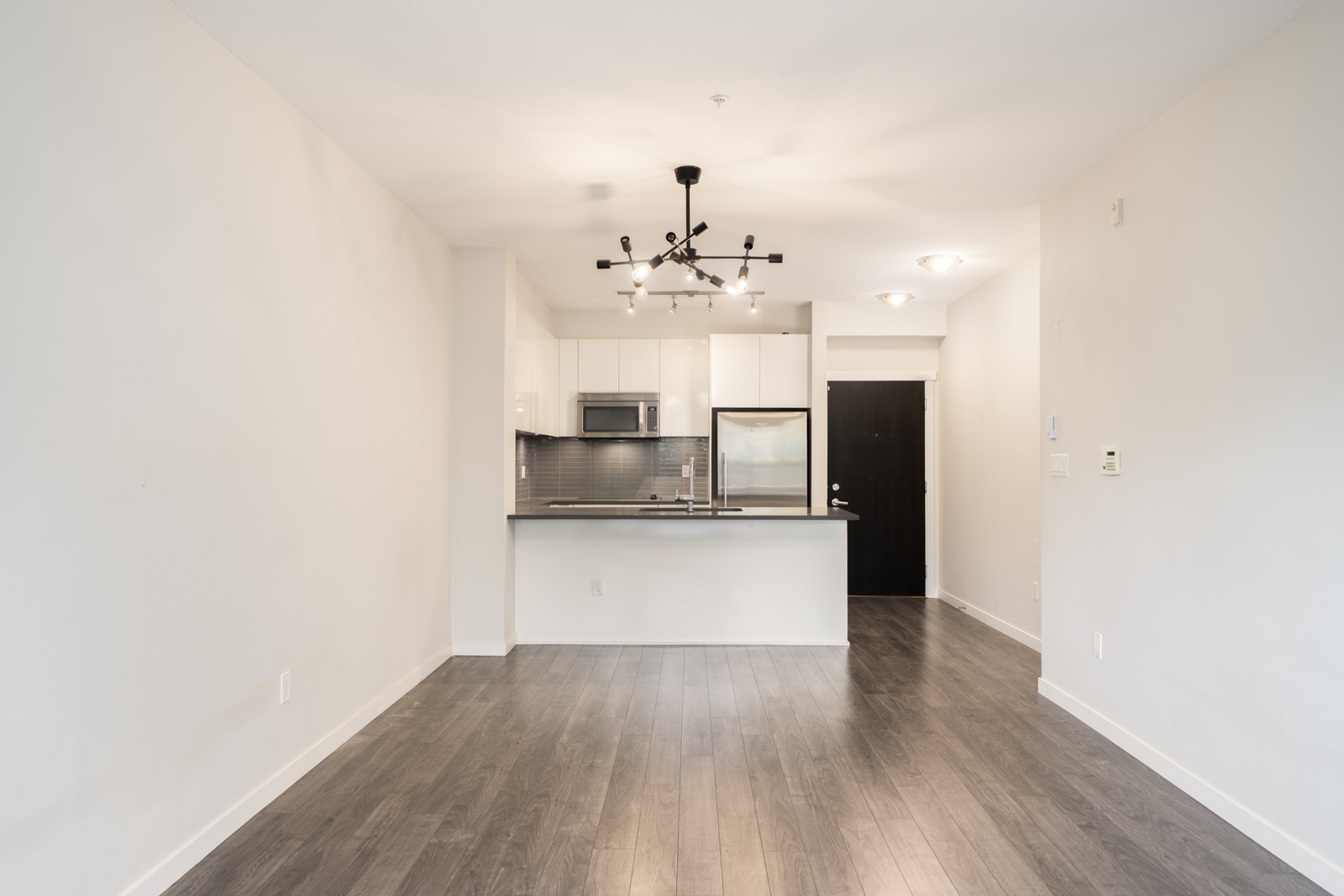 Modern North Vancouver apartment with empty living area, dark wood floors, open kitchen, white cabinets, and stainless appliances.