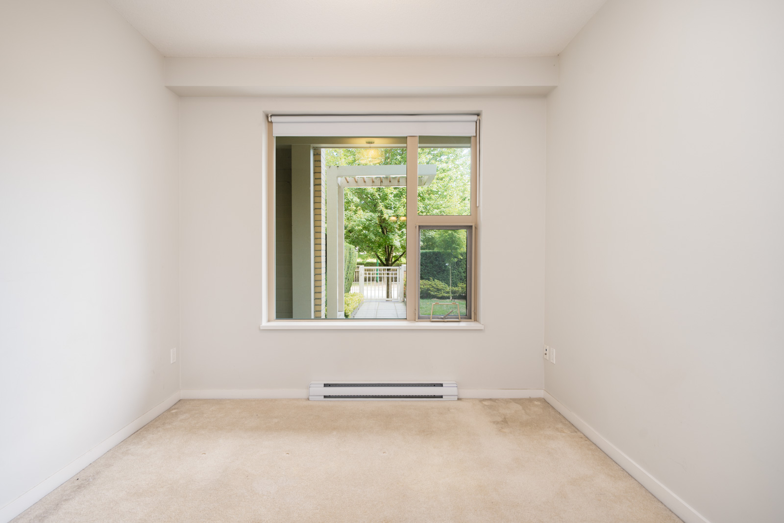 Empty room with beige carpet, white walls, and a large window facing a patio with greenery in North Vancouver on Mountain Hwy.