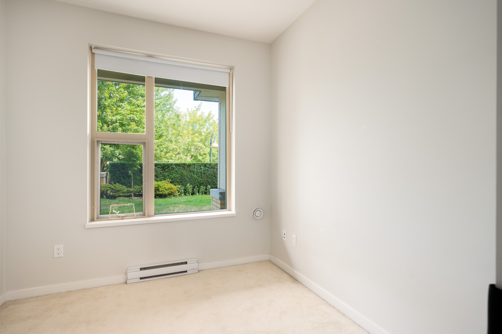 North Vancouver room with beige carpet, white walls, large window facing a green yard, and baseboard heater under window.